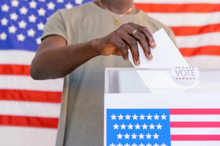 A man casting his vote into a voting box in front of an American flag at his home.