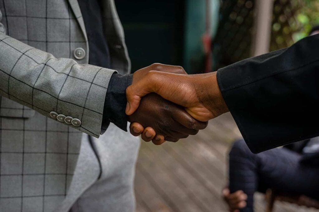 Two businessmen shaking hands in front of a group of people celebrating a win.