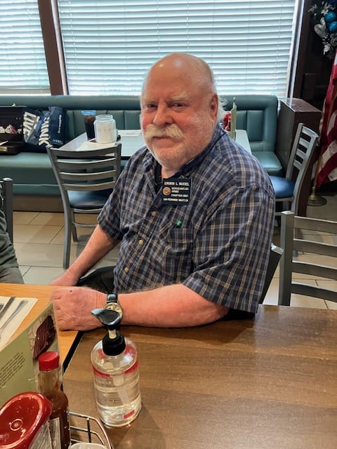 An elderly man with a mustache sitting at a table with a NarfOfficers hand sanitizer bottle in front of him.